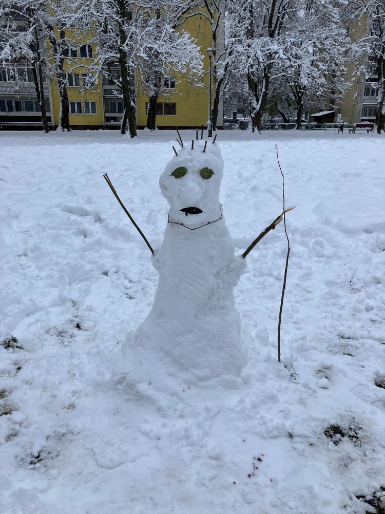 A small snowman on the snow covered grass in front of an apartment building, with stick arms, leaves for mouth and eyes and some sticks at the top for hair