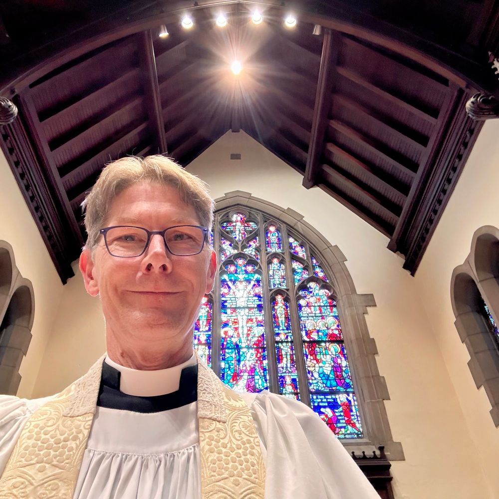 Me vested in cassock & surplice with white stole standing in front of a large and complex stained glass window — featuring Christ on the cross in the center.