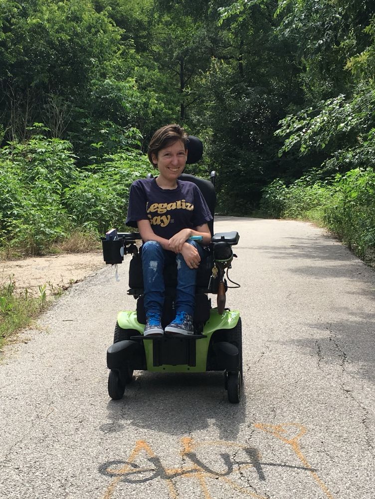 Photo of me from 10 years ago in my green power wheelchair on a trail in the woods. There is graffiti in front of me that says slut. My hair is short, I am wearing a “legalize gay” shirt and Jean leggings and converse.