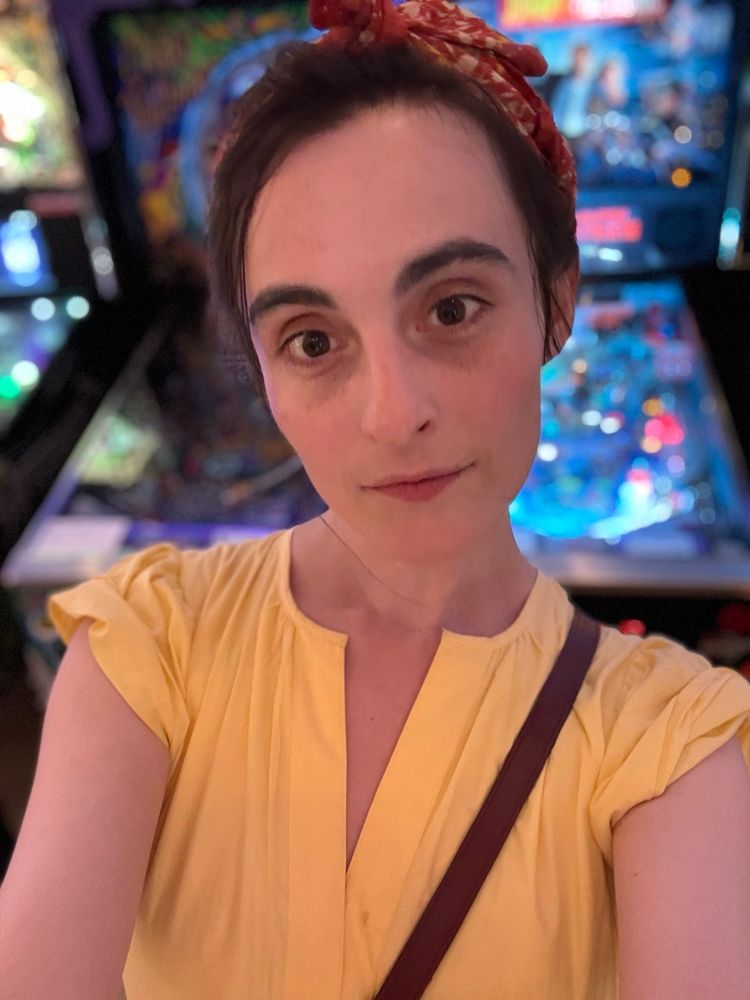 Me dark hair and yellow dress with orange and yellow leaf pattern headband standing in a pinball parlor 