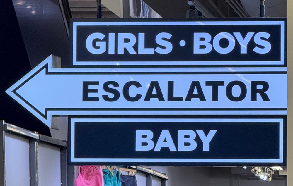 An illuminated sign hanging in Old Navy, with an arrow pointing left. It reads “Girls, Boys, Escalator, Baby.”