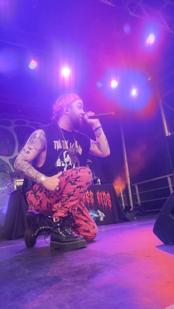 Young D kneeling on stage with the microphone to his mouth. He's wearing a ripped T-shirt type black tank top, a traditional necklace, red camo pants and platform doc Martin boots.