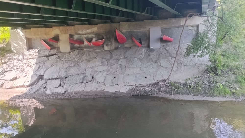 Permanent artwork under a bridge shows red canoe tips poking out haphazardly out of concrete. Large irregular shared rocked grouted with concrete form a ramp into murky waters. Green shrubbery is visible on either side of the bridge and reflected in the water.