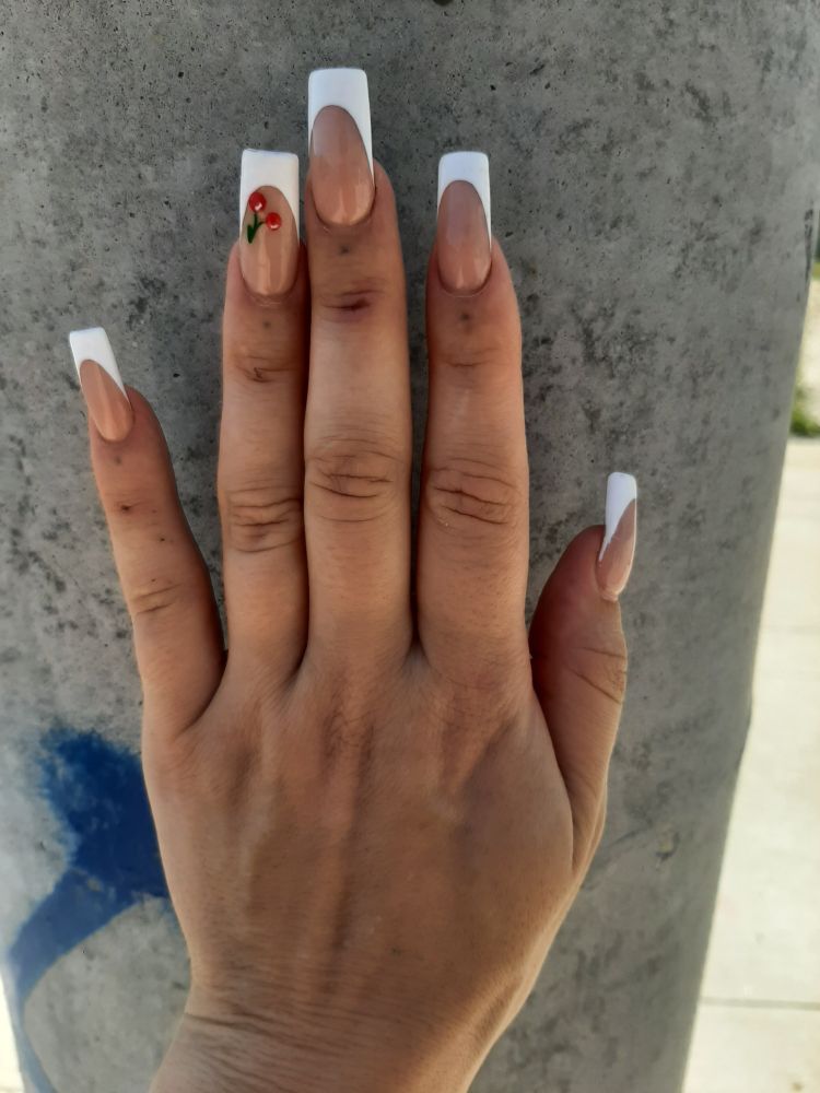 Pic of bambii's French manicure with cherry decorations on ring finger. Zir hand is tanned color and is held against a concrete streetlight post with blue graffiti visible in lower left corner