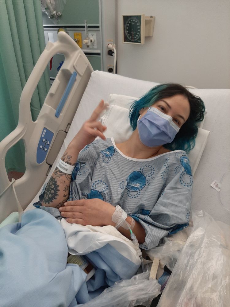 Picture of post surgery Bambii, an olive toned non binary lays in a hospital bed  with a medical gown and blanket on Zim. Zi is smiling behind Zir mask and is giving a peace sign but zi is drugged and not exactly sure what zi is doing.