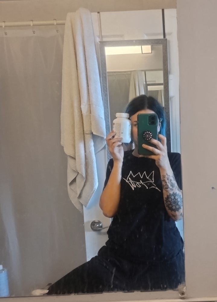 Bathroom belfie of bambii wearing a black t and sweats holding up a pill bottle the size of Zir head. The background is white toned shower curtain, wall, towel on door. 