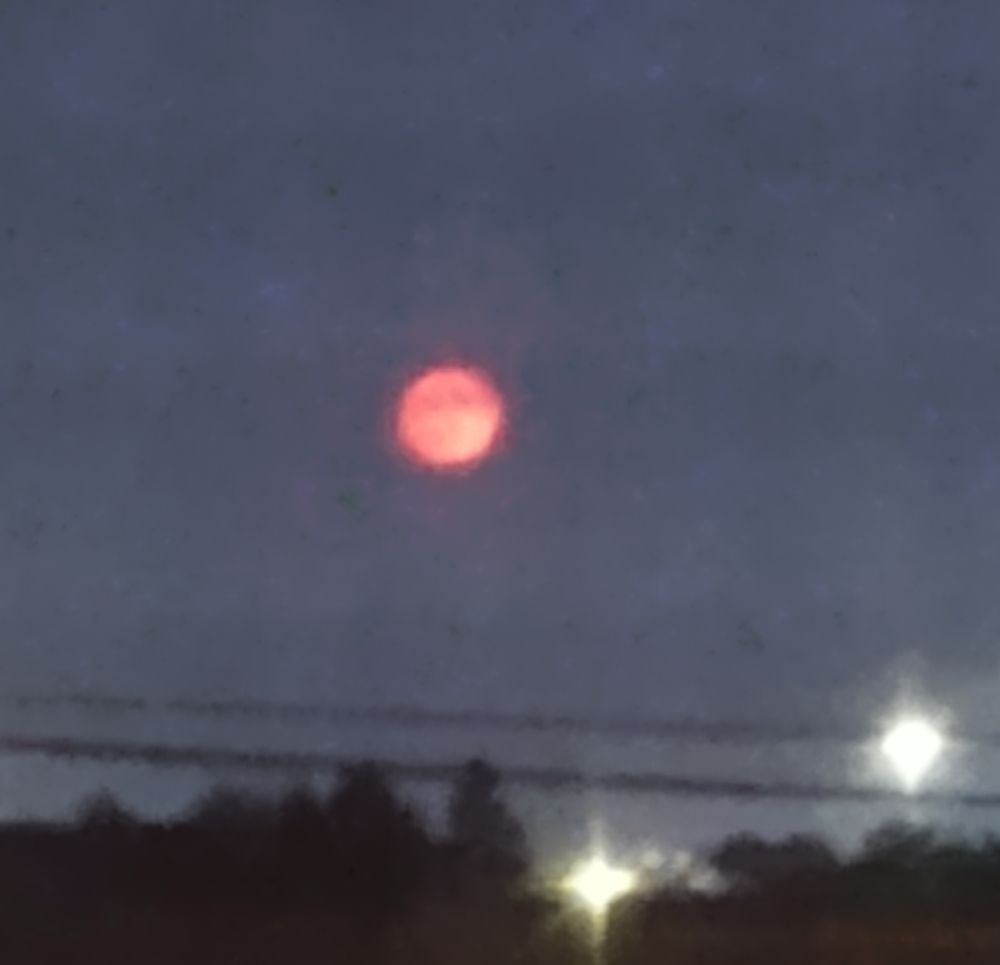 Blood moon against starless night sky top of dark trees and a couple lights at the bottom