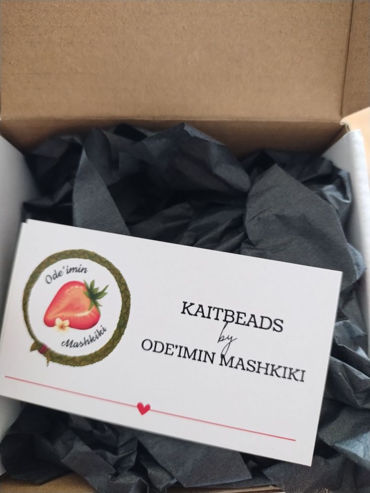 Beautiful box with black tissue paper and the business card  that reads "KaitBeads by ODE'IMIN MASHKIKI" there is a picture of a strawberry in a ring of sweetgrass on the card on the left of the writing. 