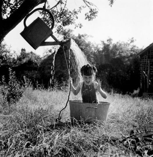Black n white photo of a young girl sitting in a metal tub, hair braided in a circle a top her head. In a grassy yard. Under a tree where a Big metal watering can is attached to a limb filled with water. The young girl pulls on the rope to douse water on her head. 