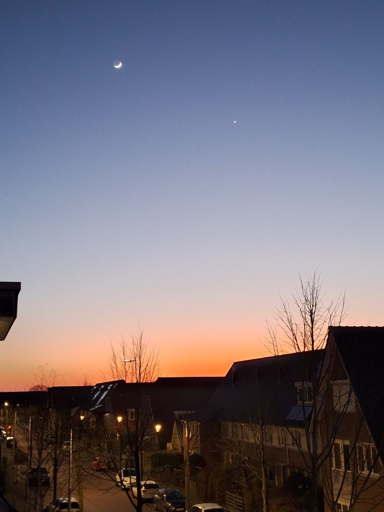 Maan, Venus en door een kale boomkruin gezien een landend vliegtuig bij een oranje zonsondergang. Vanuit zolderraam met uitzicht over straat.