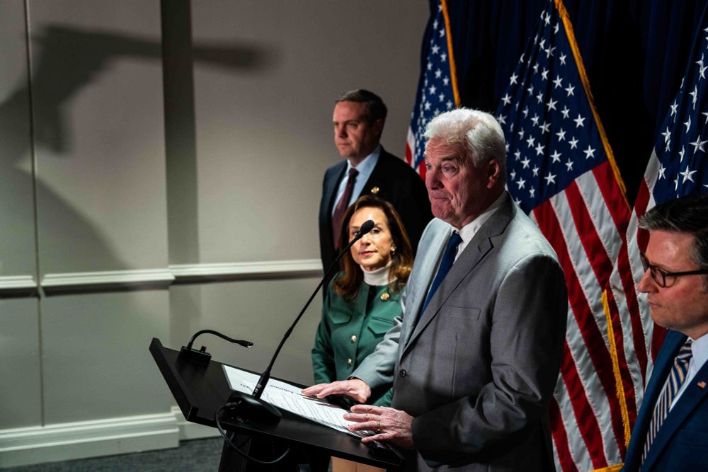 A group of individuals standing at a podium during a press conference. The central figure, an older man in a light gray suit, is speaking into a microphone, with an expression of seriousness or concern. To his right, a woman in a green outfit listens intently, while two other men in suits stand nearby, one of whom is the Speaker of the House. The background features a series of American flags, indicating an official or political setting. The lighting emphasizes the speakers, casting a shadow of the photographer holding a camera on the wall behind them.