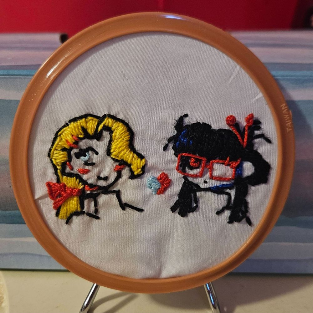 Photograph; Embroidery of two women, one with a low blond pony tail and the other with black hair in a bun with red hair sticks and glasses.