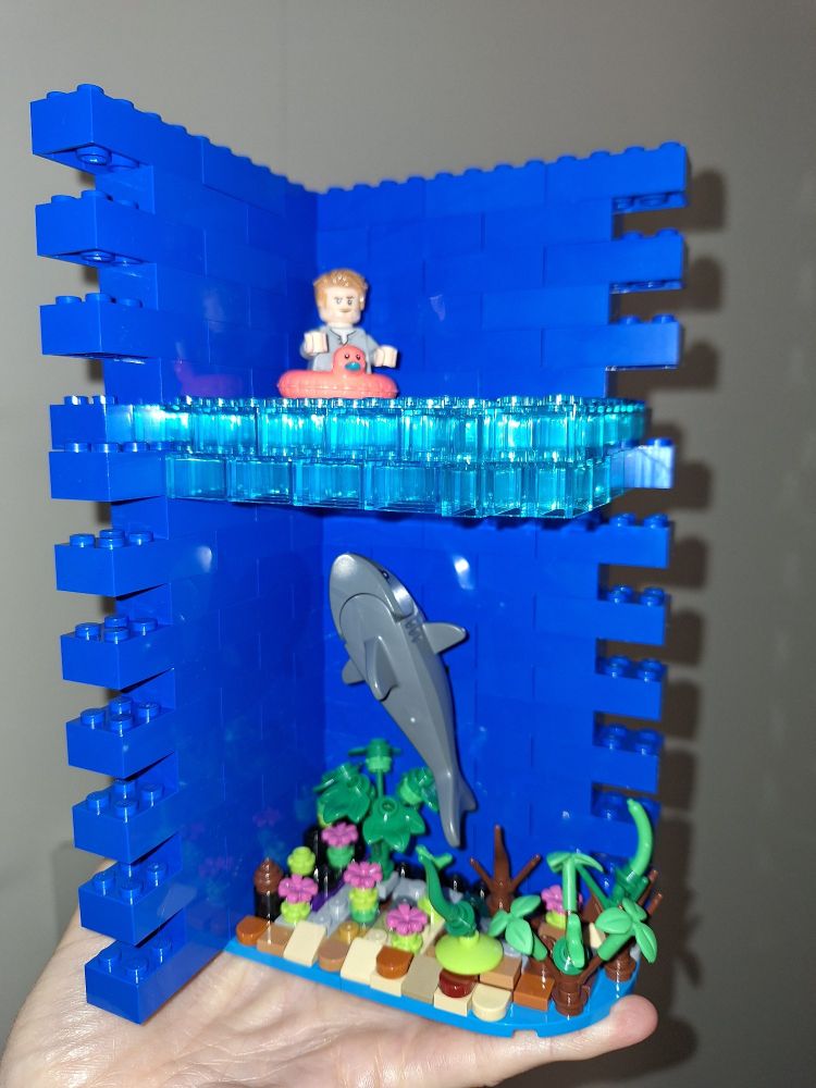 Kleines Diorama aus Lego gebaut. 
Blaue Steine im Hintergrund für Meerwasser und Himmel.
Transparente Steine stellen die Wasseroberfläche dar.
Der Meeresboden ist mit bräunlichen und grauen Steinen angedeutet. Auf dem Grund sind einige Pflanzen.
Ein Legomännchen mit Schwimmring treibt auf der Oberfläche.
Und darunter kommt ein Hai angeschwommen.
