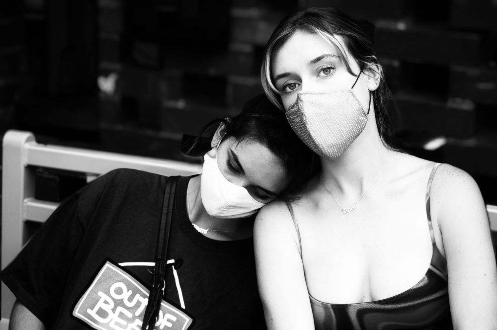 Two women sitting on a bench. One looking into the camera, the other resting her head on the first’s shoulder. 