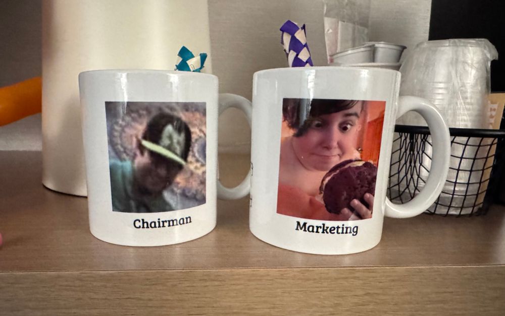 A pair of mugs with terrible photos of the chairman and marketing lead of Furcationland on a shelf.