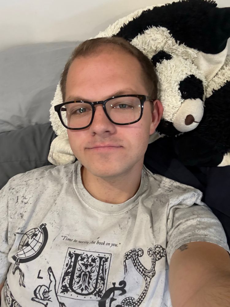 Will, a gay, laying down taking a selfie in a white/light grey tshirt 