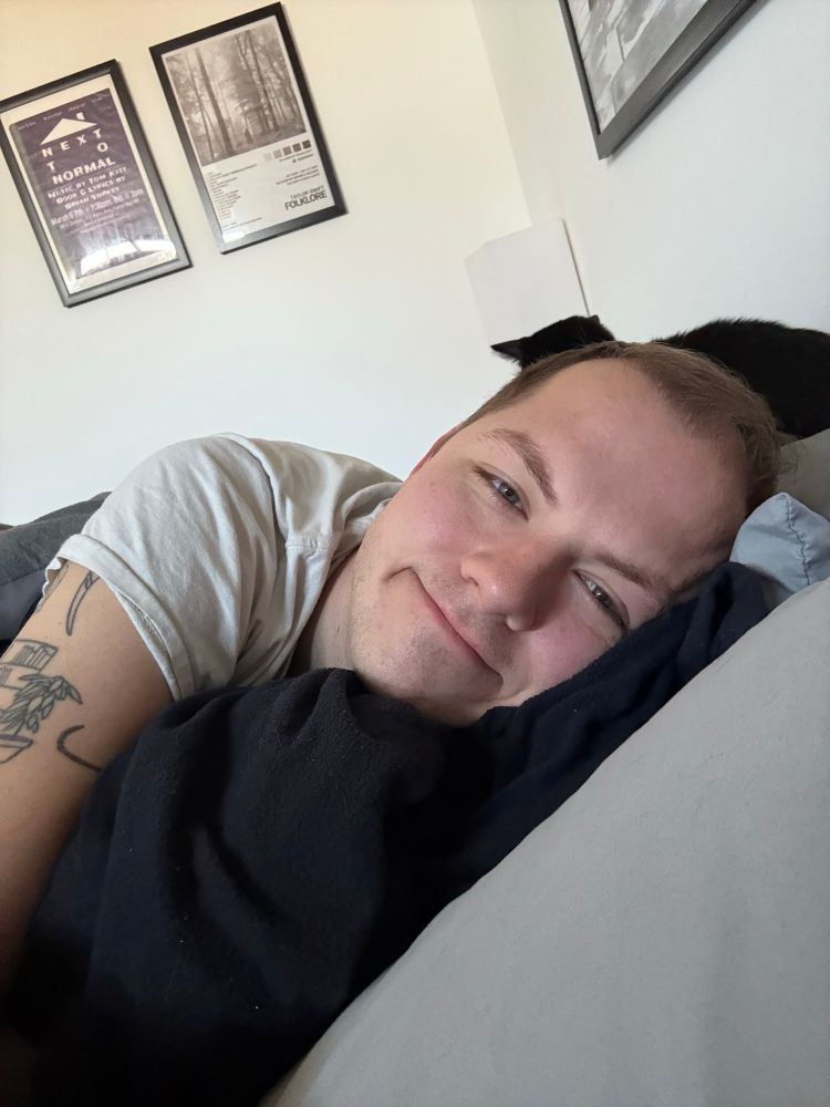 Will, a gay, laying in a bed. A cat is perched on their pillow 