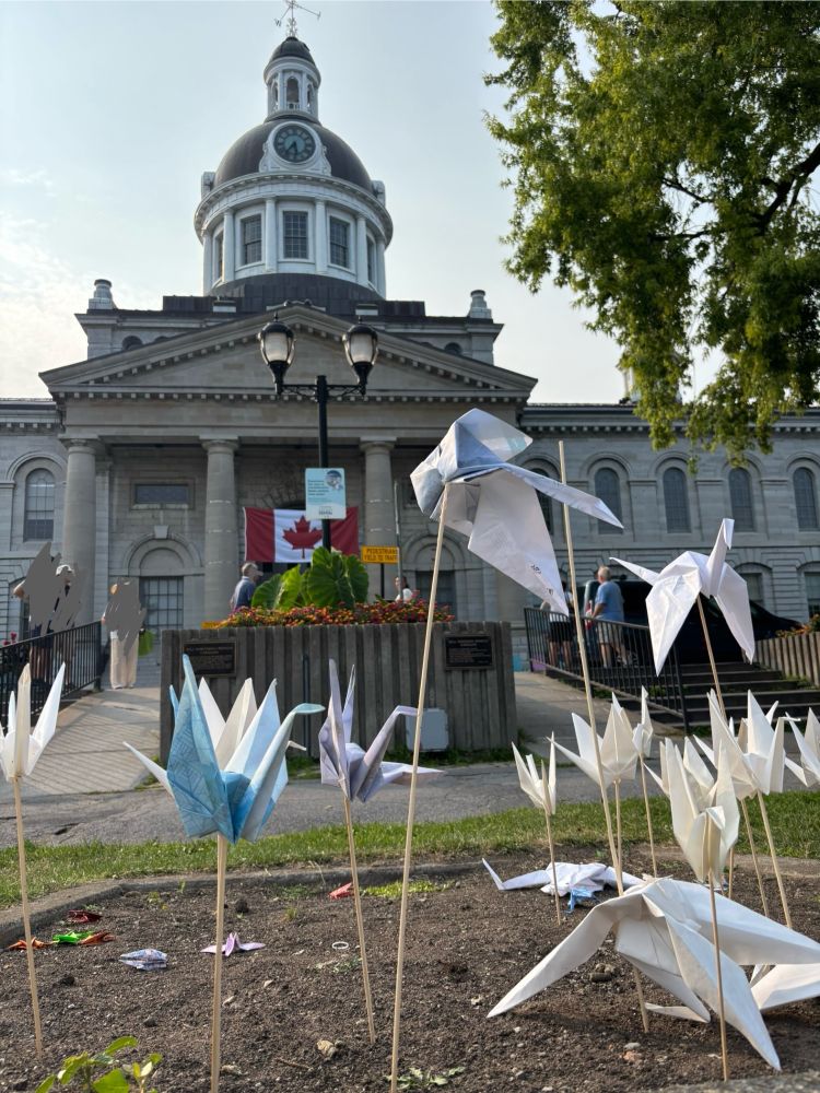 Grey stone buiding in background, with domed roof & Canada flag. Foreground has paper cranes on sticks 