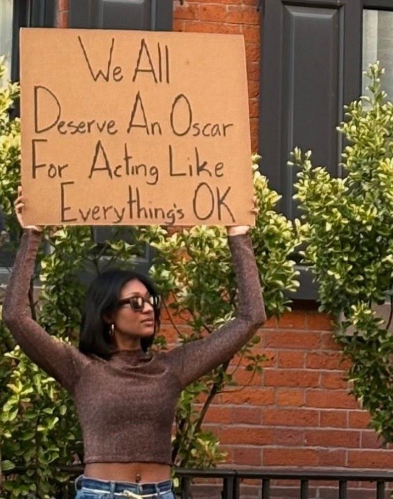 A person with black shoulder length hair & sunglasses holding a sign that reads “We all deserve an Oscar for acting like everything’s OK”