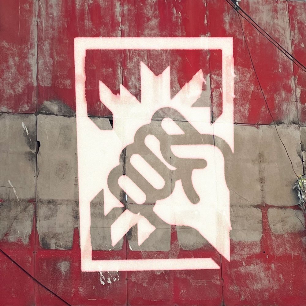 Im Hintergrund ist eine Metallwand zu sehen, welche einen weinroten Anstrich bekommen hat und bereis verblasst. Es hängen einzelne Kabel an der Metallwand entlang. Im Vordergrund ist mittig zentriert ein Piktogram einer Hand in weiß, die ein Fenster einschlägt.