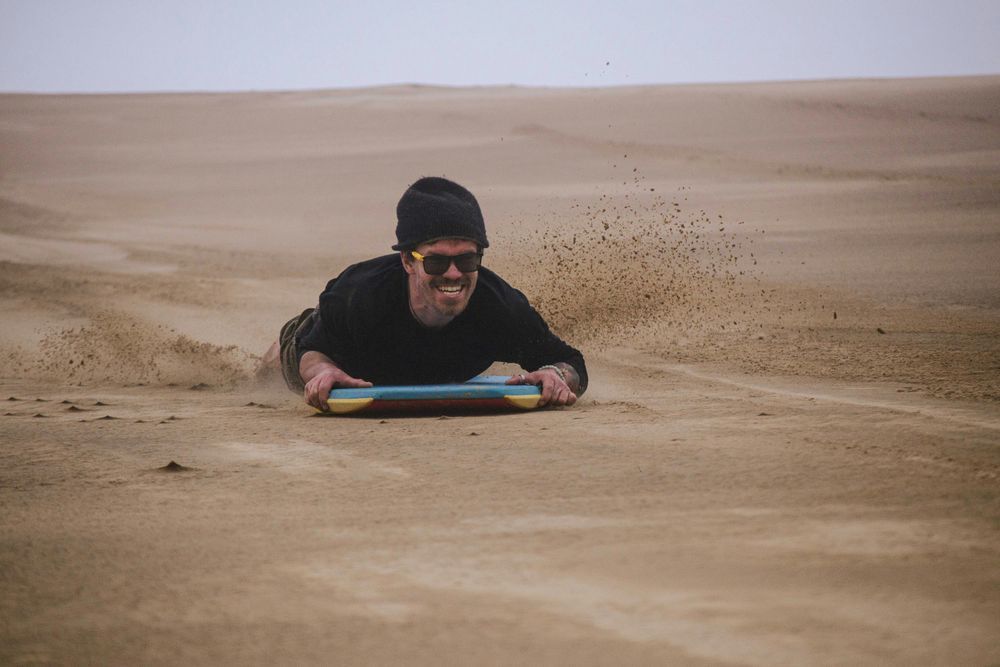 A guy having a great time sandboarding. He's prone on a sandboard, grinning as the sand scatters behind him.