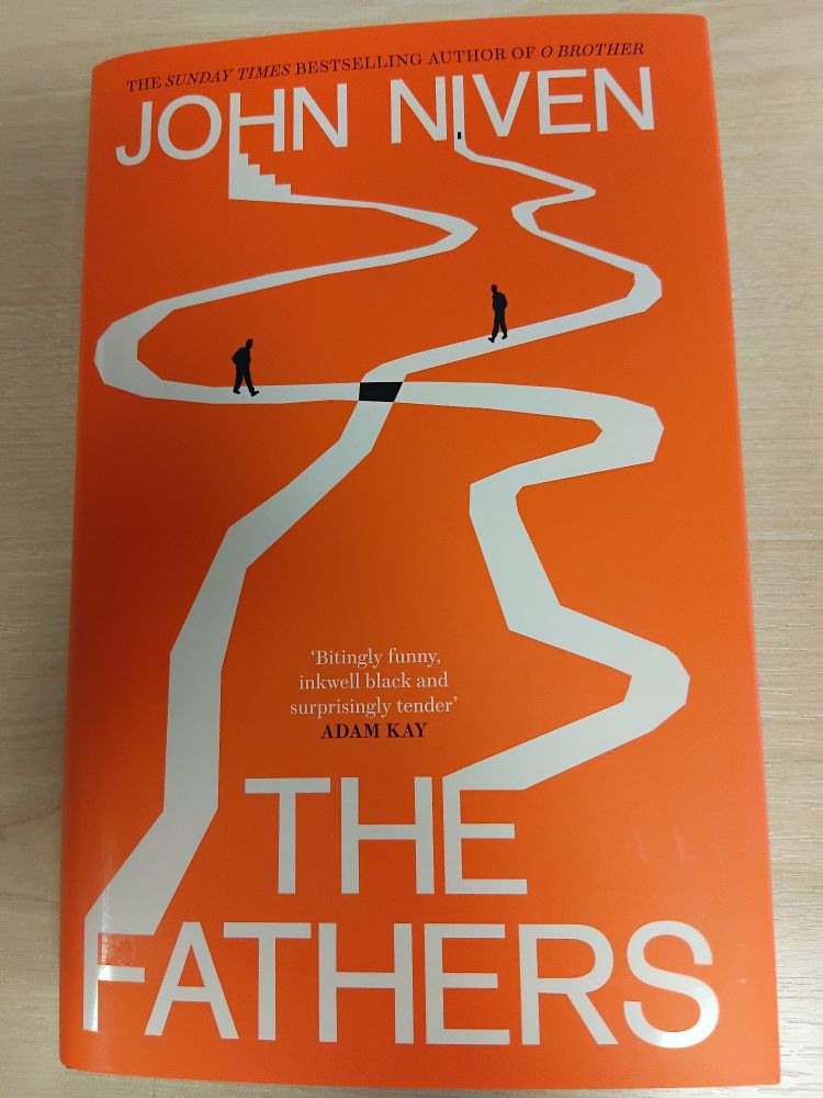 Cover of John Niven's new novel The Fathers. It has an orange background with an illustration of two white paths crossing over each other. 2 men walk the paths and will meet where the paths intersect.