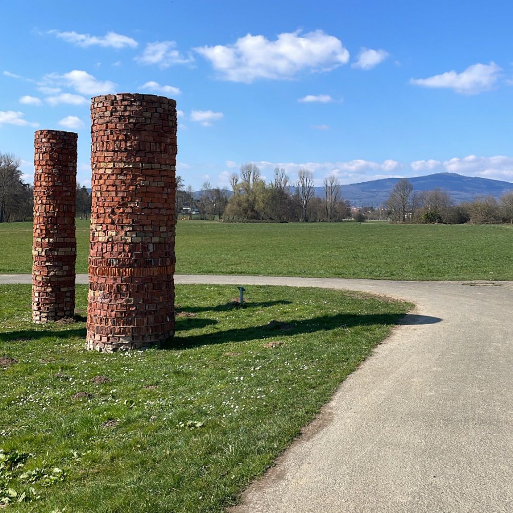 Skulptur aus Stein auf Wiese. 2 Säulen am Wegrand im Skulturenpark vor blauem Himmel und dem Taunus im Hintergrund. 