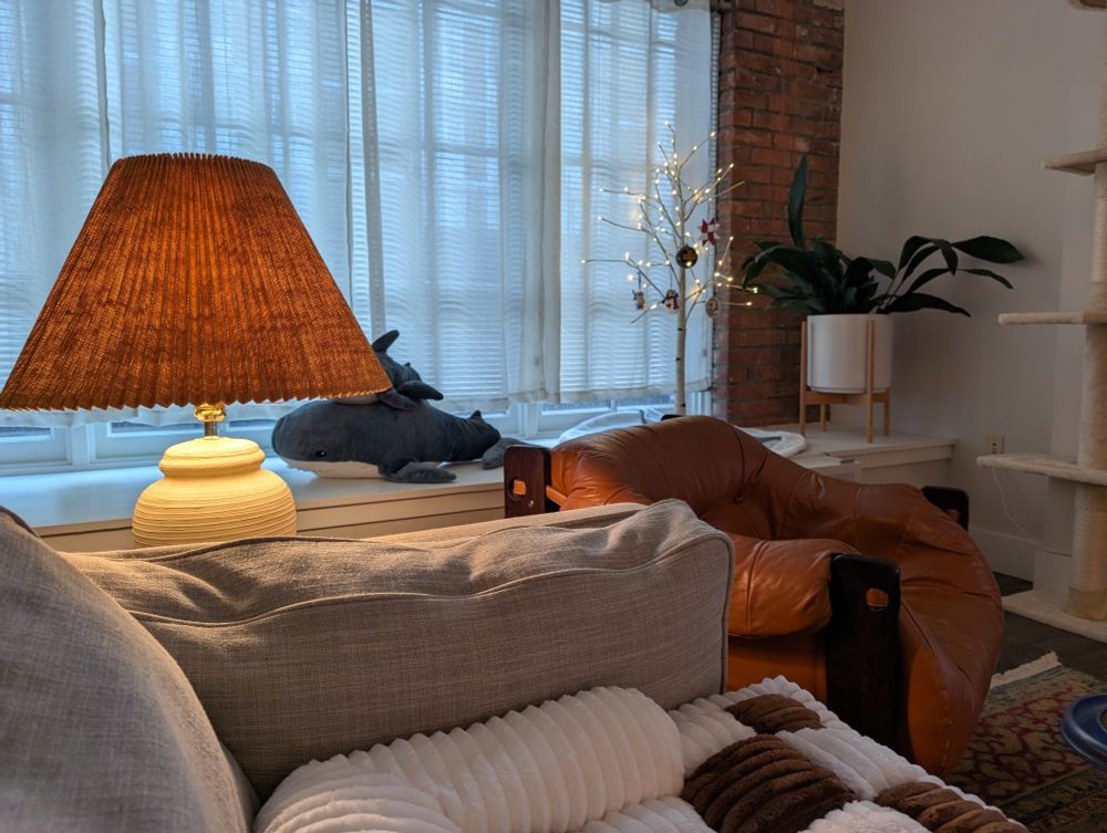 Image of a living room. Visible in the foreground is a grey sofa, a warm-colored vintage beehive lamp, and an orange leather lounge chair. In the background is a cast iron plant, a Christmas tree, some IKEA blahaj plushie sharks, and a large sunny window surrounded by exposed brick.