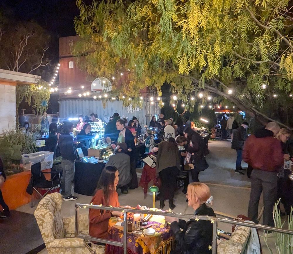 An outdoor market. It is evening, and the market tables are lit by string lights. Many people look at items and speak with vendors.