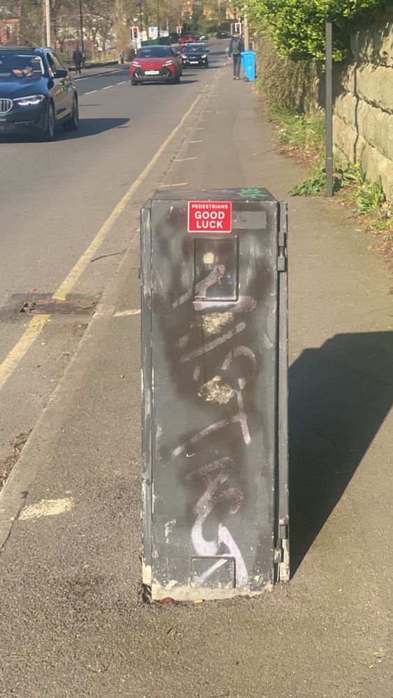 Sunny Talbot Street, Sheffield, a black metal box in the middle of a pavement with cars driving past. A red sticker on it says “pedestrians good luck”
