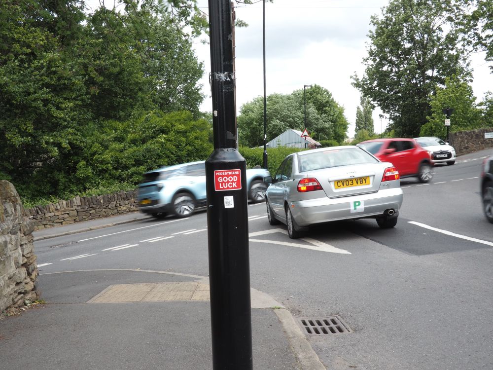 Cars moving in all directions at a cross roads jucntion with a black post with red sticker saying “pedestrians good luck”