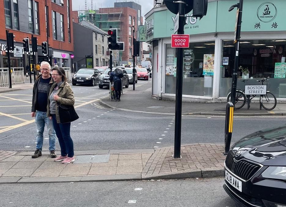 A couple of people stand on a small island between carriageways with no pedestrian crossings while a car drives from the side of them. A pole in the island has a red sign saying “pedestrians good luck”
