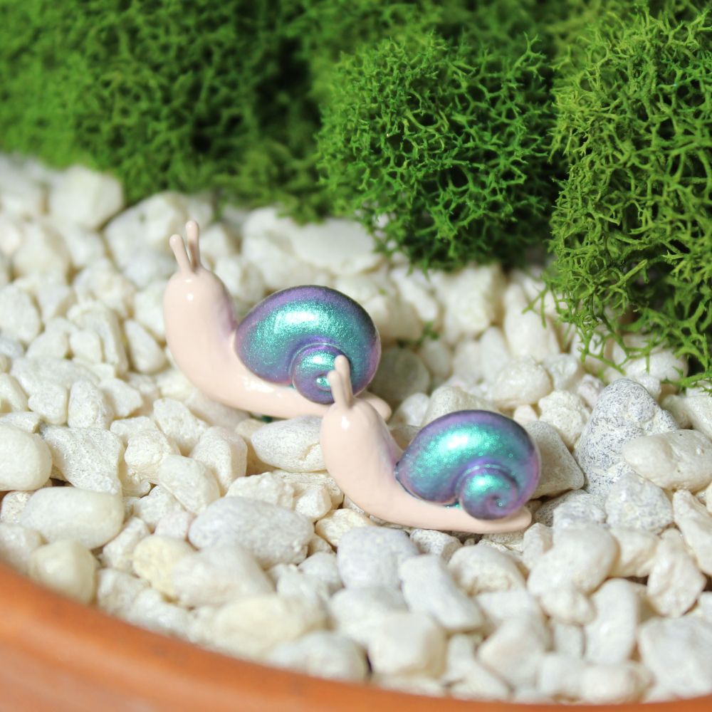 A pair of miniature snail figurines with pastel blue and purple shells sit in a mossy planter