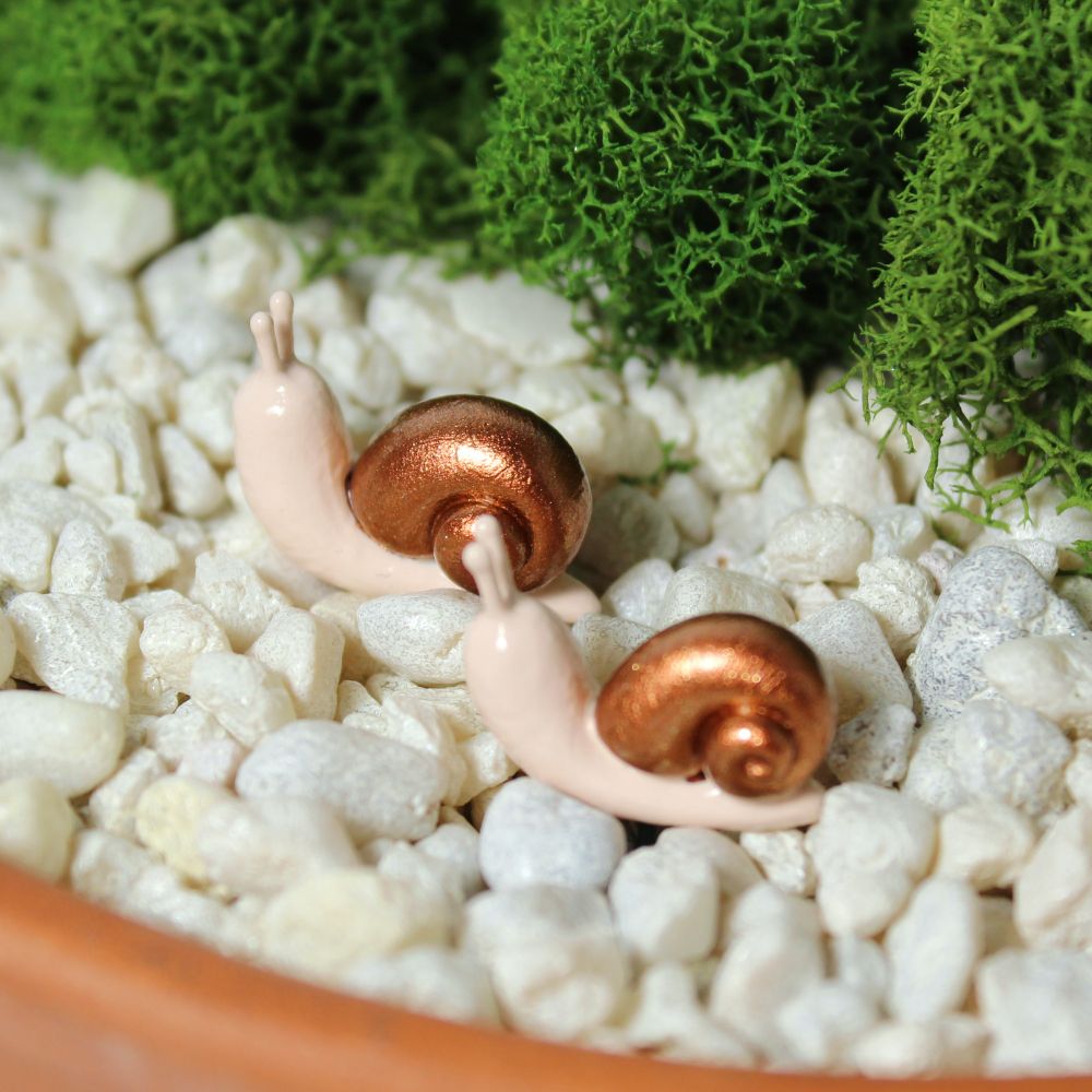 A pair of miniature snail figurines with sparkly copper shells sit in a mossy planter
