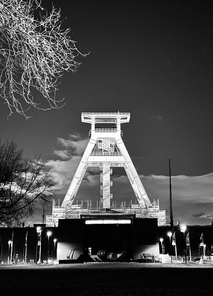 Schwarz-weißes Foto eines hellen Förderturms vor schwarzem Nachthimmel mit ein paar Wolken, unterhalb des Förderturms sind einige Straßenlaternen vor dem dunklen Gebäude zu sehen, links oben ragen einige angestrahlte Zweige ins Bild
