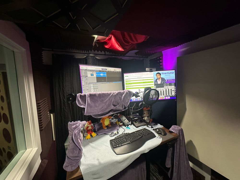 The inside of a vocal recording booth. The two display monitors show a Logic Pro X DAW session and a split screen of a dummy script and a video of Kiran Kumar’s animation demo, respectively. 