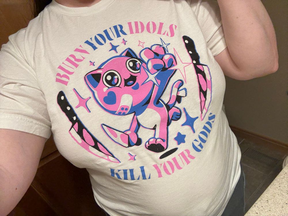 A white t-shirt with a pink and blue cat holding up a V victory hand while also holding a knife. Two more knives sit on both sides of the cat. Top text reads “Burn on your idols.” Bottom text reads “Kill your gods.”