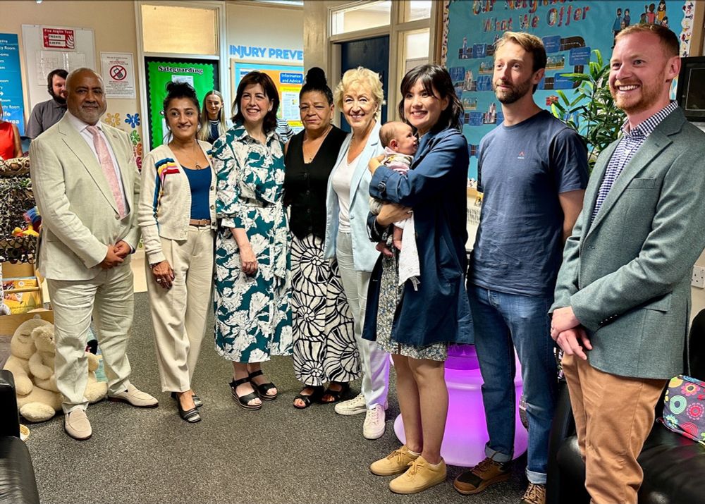 A group of people are stood smiling in the family centre, with Lucy Powell and Dame Andrea Leadsom.