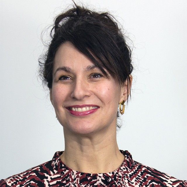 A woman with dark curly hair tied up wearing gold hoop earrings and a patterned red, black, and white top against a plain light background.