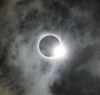 A total solar eclipse.  Whispy clouds cover a dark circle with a ring of bright light around it.  On one side is an especially bright light, like a diamond on a ring.