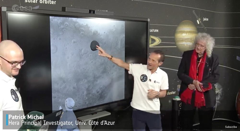 HERA principal investigator Patrick Michel pointing at a TV screen showing Mars' moon Deimos (black blob) over the surface of Mars (cratered).  Rock legend with long rock hair, Brian May, standing the background looking on.