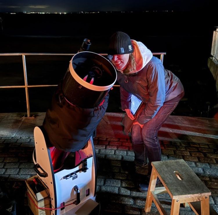 A woman (me) smiling, with black woolly hat and lots of layers, peering through a relatively large telescope.