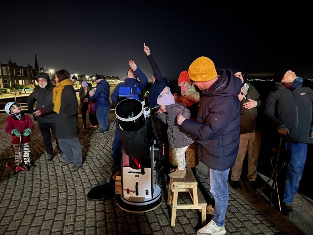 Lots of people looking up, point at the night sky.
A cut little one is standing on a stool looking through the telescope.