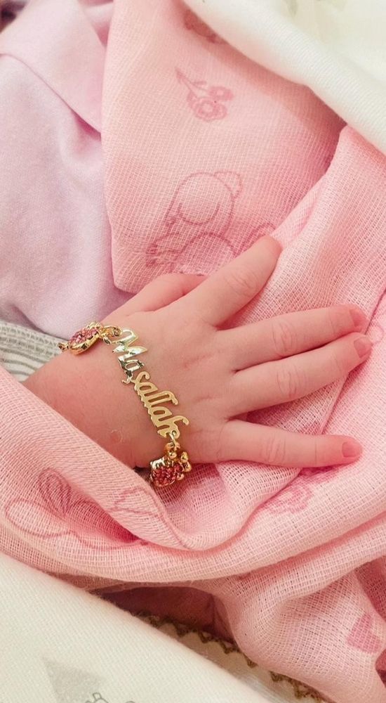 An infant's righ4 hand wearing a tiny charm bracelet that says Masallah. Pink and white background.