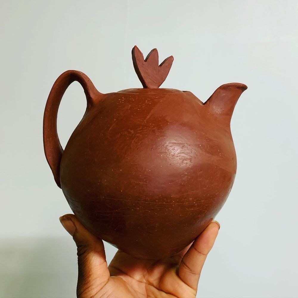 My hand holding a small, unfired teapot at the leather hard phase, so the clay is still a rich, brown color with a slight sheen. The pot features a stylishly curved handle, a small spout and a lid with a finial that sort of resembles the comb on a rooster’s head or perhaps an abstract flower. 