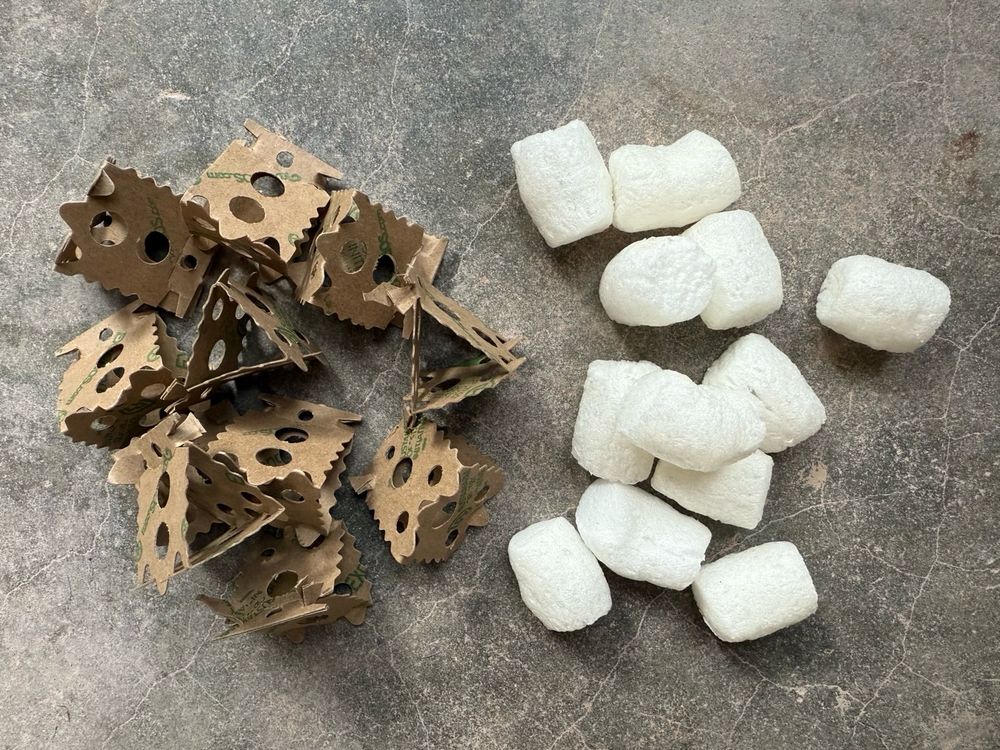A photo looking down at two piles of materials. On the left, a handful of brown paper pyramid-shaped packing materials. On the right, white puffy foam peanuts made from biodegradable corn starch. 