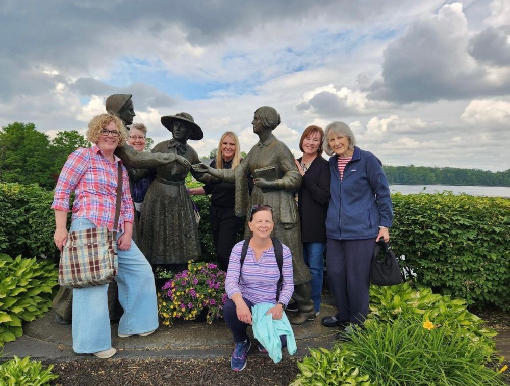 A group of 6 historians pose with a statue of SB Anthony, AJ Bloomer, and EC Stanton in Seneca Falls, NY.