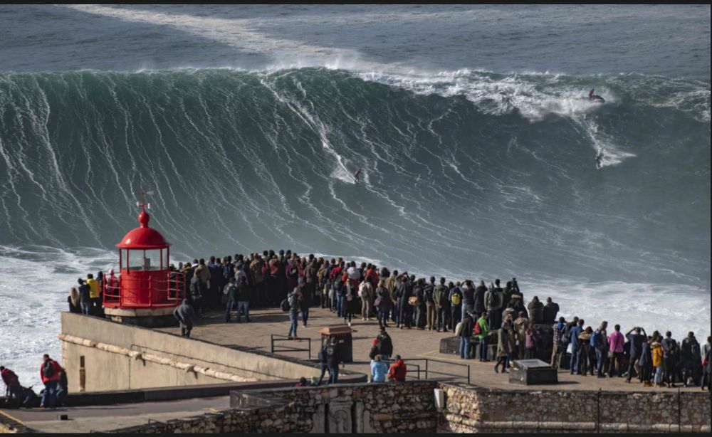 Picture of a surfer riding a huge wave with spectators standing on a cliff near a lighthouse. 