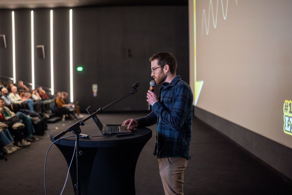 Julien Sulpis, focused and looking at his laptop, holds a microphone and clicks on his Macbook on stage during his presentation at the first LyonJS conference.
