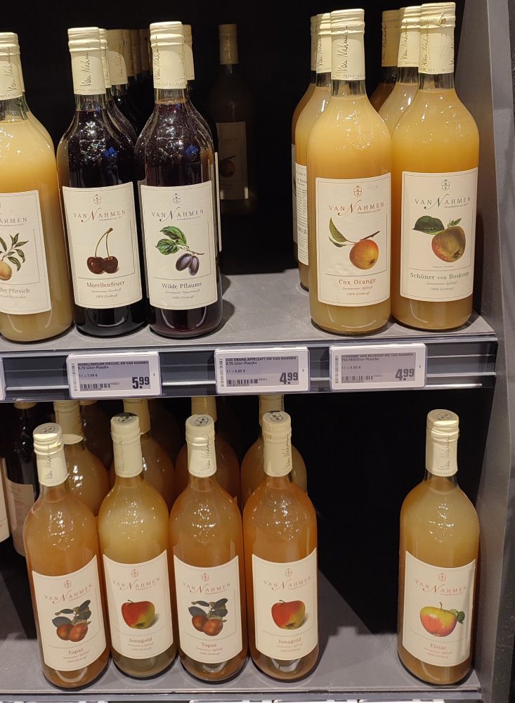 Glass bottles with white labels each with a color painting style illustration of different types of apples including Topaz, Jonagold, Orange Cox and Boskoop as well as other juices. The apple juices vary in color between yellow and orange. The price is between 4 and 5 € 
